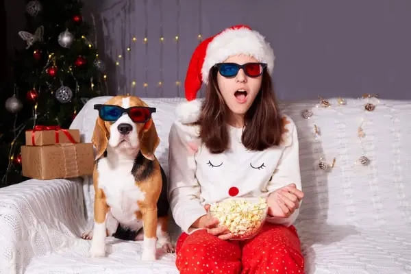 A girl in a Santa Claus hat and 3D glasses is sitting on the couch with a Beagle dog, watching a Christmas movie.