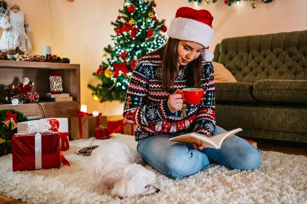 Woman reading book and drinking hot chocolate during Christmas with her Maltese dog.