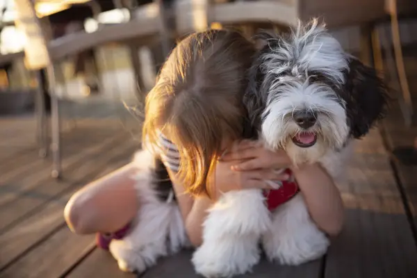 Child hugging young Havanese puppy next to an outdoor dining area.